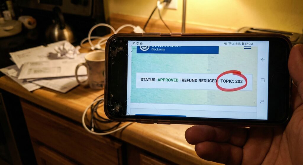 A close-up of a smartphone screen displaying the "Tax Topic 203" warning on the IRS Where's My Refund tool.
