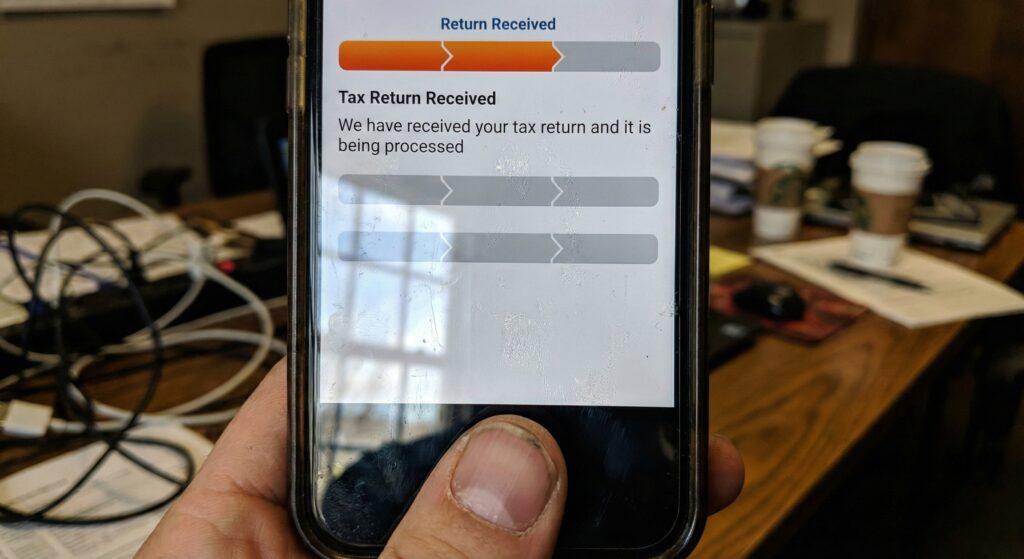 A frustrated person holding a smartphone showing the IRS "Where's My Refund" app stuck on the first bar "Return Received".